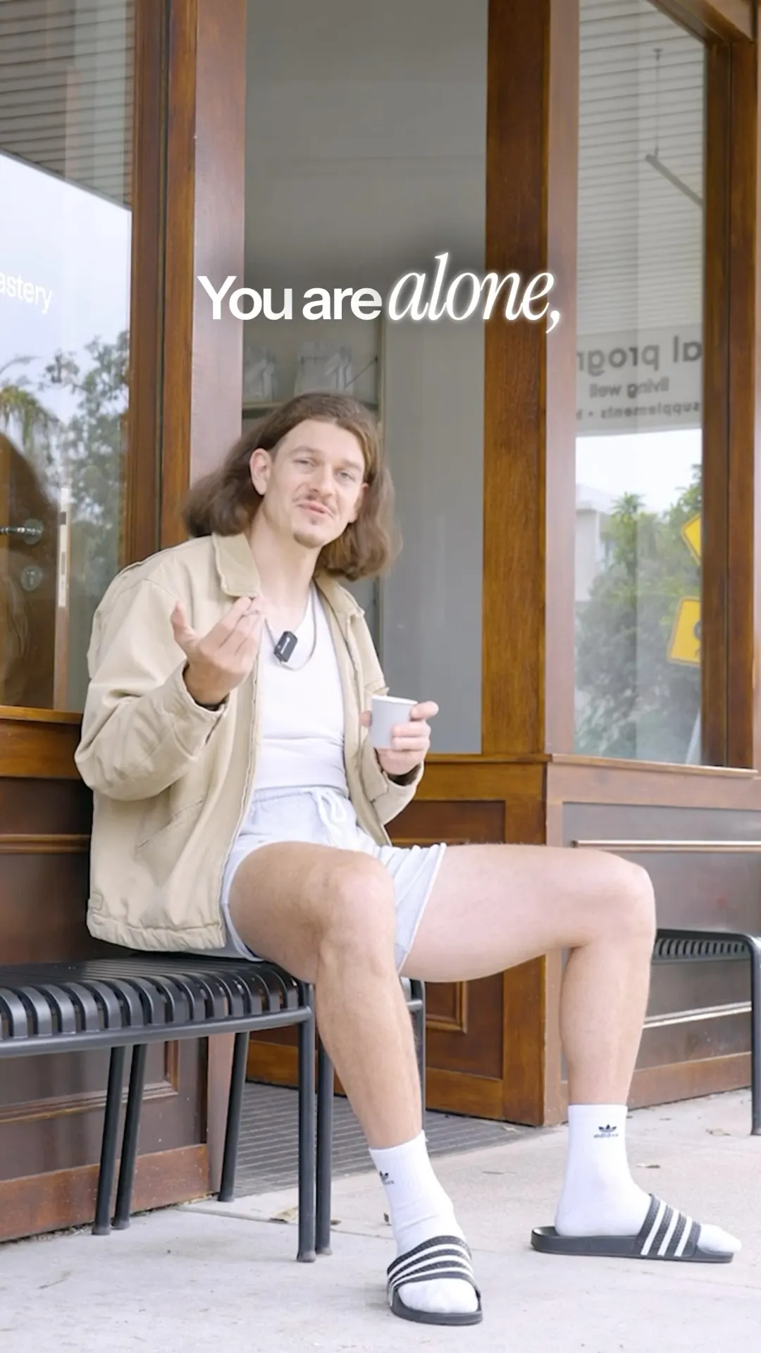 A man in a beige jacket and shorts sitting on a bench outside a shopfront, holding a small takeaway coffee cup.