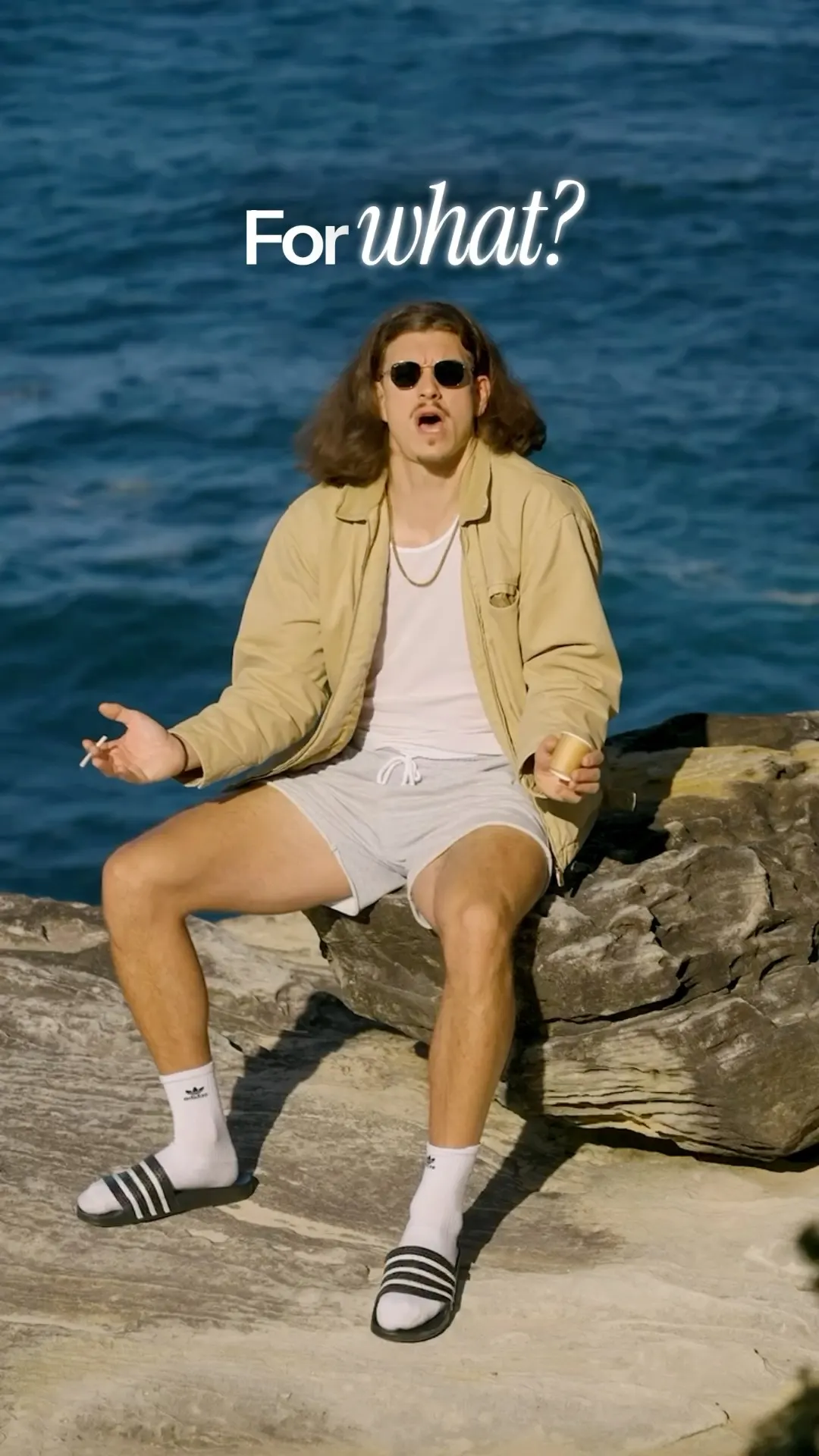A man in a beige jacket and shorts sitting on a rock shelf beside deep blue water, hands open mid-sentence.