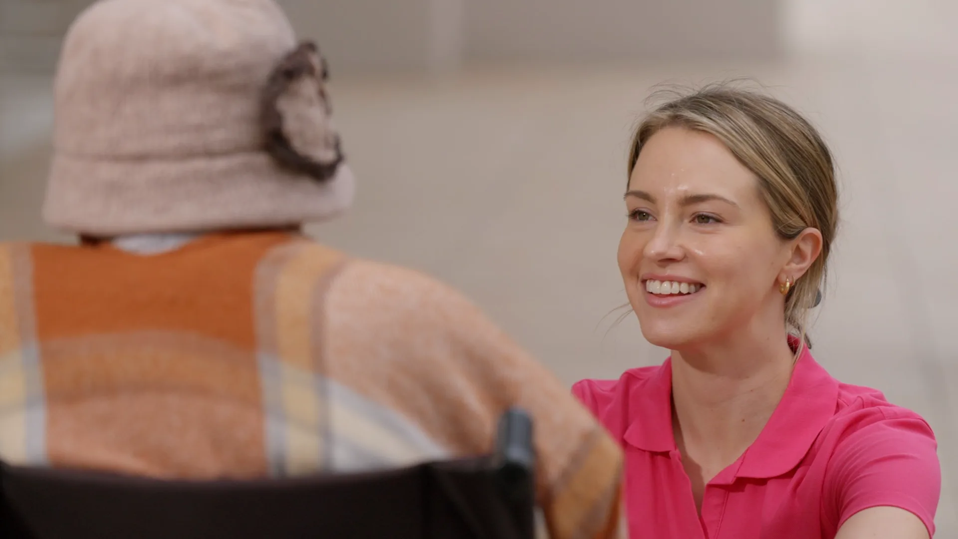 A woman in a pink polo shirt smiling as she talks with a seated person wearing a hat, indoors.