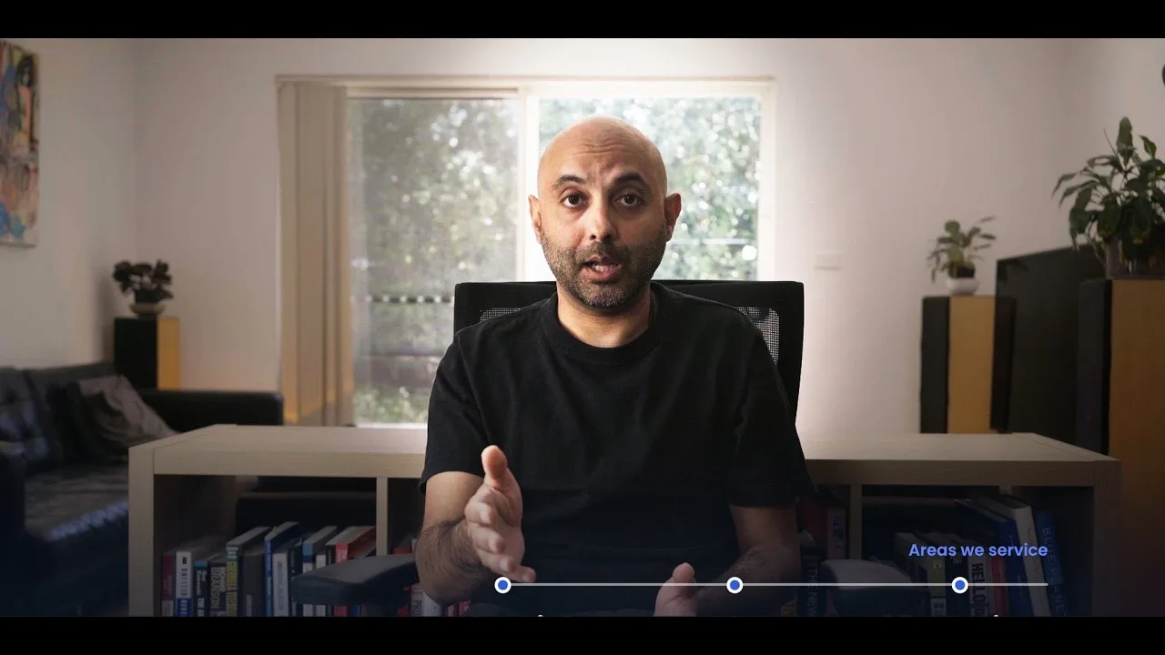 A man in a black t-shirt speaking to camera in a home office, with a window and bookshelf behind him.