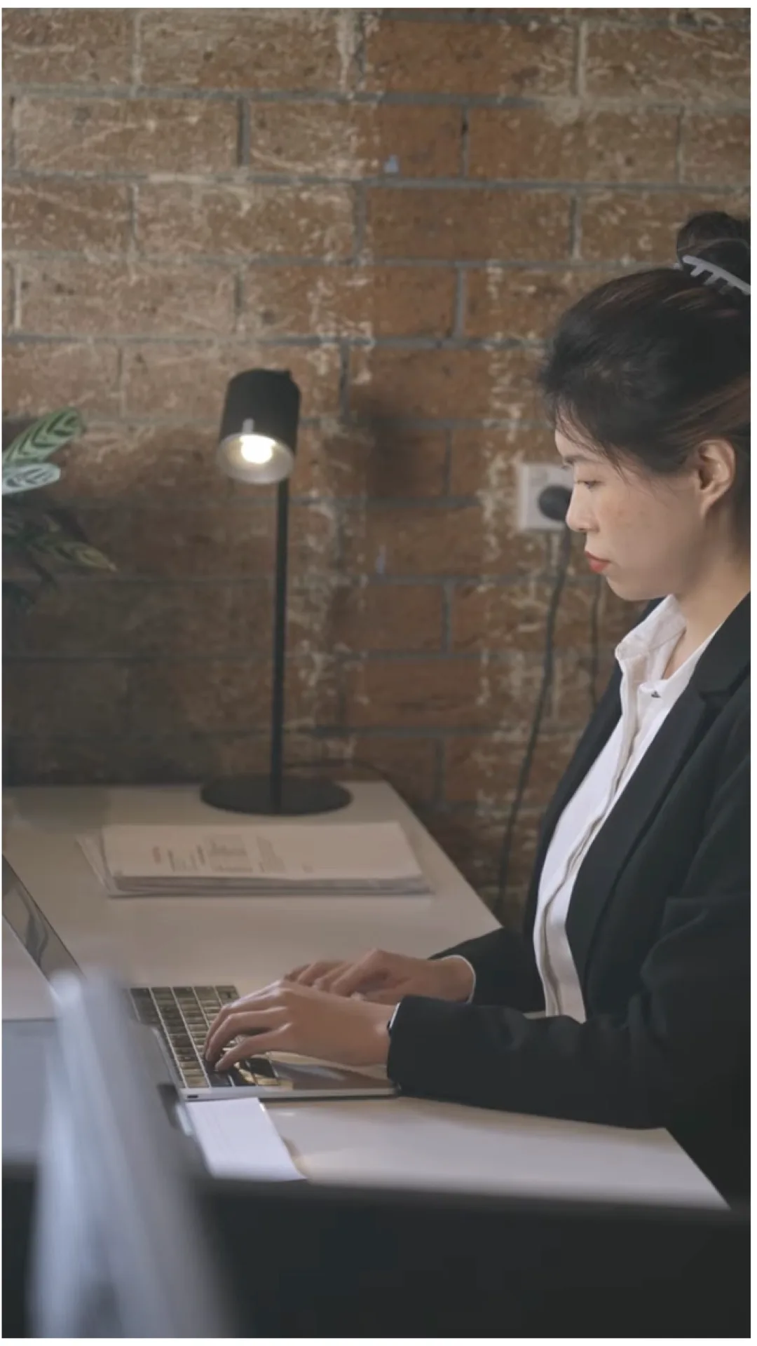 A person in a dark blazer typing on a laptop at a white desk against a brick wall, lit by a desk lamp.