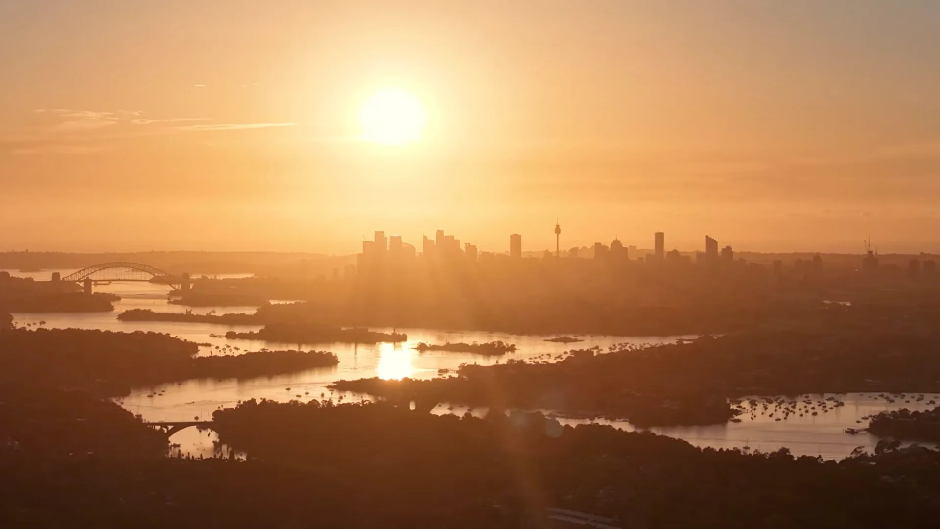 Wide aerial view of Sydney at sunrise, the sun above the city with the Harbour Bridge and the river below.